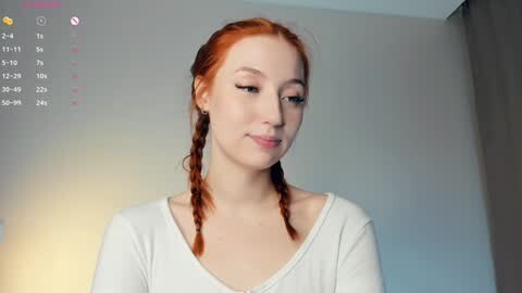 Media: Video of a fair-skinned young woman with long, red braided hair, wearing a white off-shoulder top, standing against a light-colored wall.