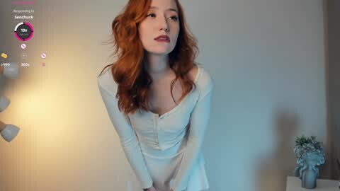 Media: Video of a fair-skinned redhead woman in a white dress, standing indoors against a plain wall with a potted plant and scattered photos.