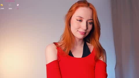 Media: Video of a fair-skinned, red-haired woman in a red off-shoulder top, smiling softly. Background is a plain, light-colored wall with a blurred curtain.