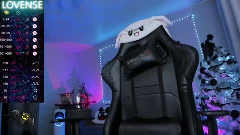 Media: Video of a black, high-backed gaming chair with a white, smiling cat head cushion. Background includes a festive Christmas tree and multicolored lights, with a digital overlay displaying a chat window.
