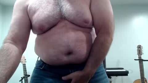 Media: A video of a shirtless, middle-aged man with a hairy chest and belly, wearing blue jeans, standing in a room with guitars and an amplifier in the background.