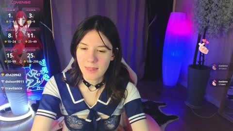 Media: A young woman with fair skin, long black hair, and a white and blue sailor-themed outfit, wearing a choker, sits in a dimly lit room with purple and blue lights. She's in a video game stream.