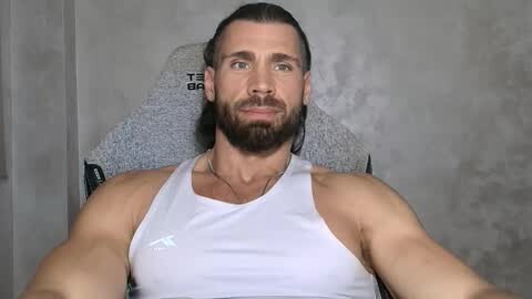 Media: Video of a muscular, bearded man with a white tank top, seated on a gray chair with a textured cushion, against a plain gray wall background.