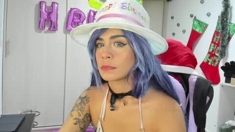 Media: Video of a woman with light skin, blue hair, and a white hat with \"Happy Halloween\" in green. She wears a black choker and a purple bikini top, sitting in a gaming chair with festive decorations in the background.