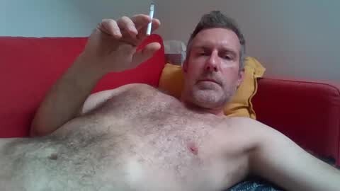 Media: Video of a middle-aged man with a stubble beard, lying shirtless on a red couch, holding a thermometer, wearing a yellow pillow.