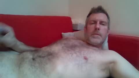 Media: A video of a middle-aged man with short brown hair, light skin, and a hairy chest, lying on a red couch, slightly out of focus, in a dimly lit room.