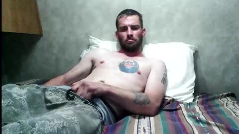 Media: Video of a shirtless, fair-skinned man with short hair, tattoos, and a blue and red circular tattoo on his chest, lying on a bed with a striped blanket, in a dimly lit room with a wooden nightstand.