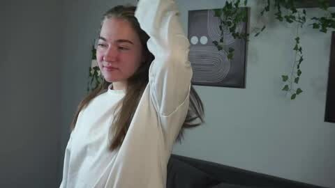 Media: Video of a young Caucasian woman with light skin and straight, light brown hair, wearing a white long-sleeve shirt, stretching her arms upward. Background features a gray couch, wall art with abstract patterns, and green plants.