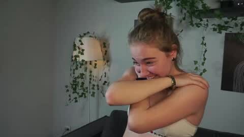 Media: Video of a young woman with fair skin, brown hair in a messy bun, and a small tattoo on her right arm. She's laughing, hugging her arms, and wearing a towel. Background features a green plant and a lamp.