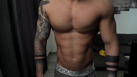 Media: Video of a shirtless, muscular man with tan skin, wearing Calvin Klein underwear, black arm tattoos, and a black wristband. Background features a dark sofa and cityscape artwork.
