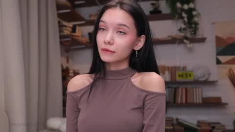 Video of an East Asian woman with straight black hair, fair skin, and a slender build, wearing a brown off-shoulder top, standing indoors amidst shelves filled with books and plants.
