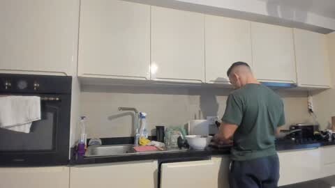 Media: Video of a muscular, bald man in a green t-shirt, washing dishes in a modern kitchen with white cabinets and black countertops.