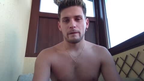 Media: Video of a shirtless, fit white man with short, light brown hair and a trimmed beard, wearing a necklace, sitting indoors with beige walls and a wooden window.