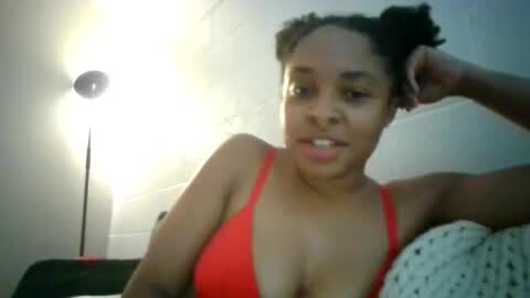 Media: Video of a young woman with medium brown skin, curly hair in pigtails, wearing a red bra, leaning on a white pillow with a polka-dot pattern, against a white brick wall.