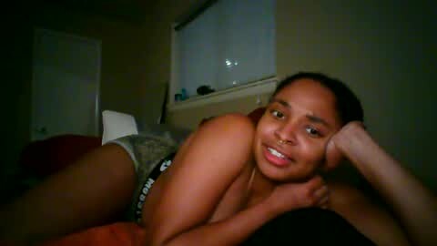 Media: Video of a young, dark-skinned woman with medium-sized breasts, wearing grey shorts and a black bra, lying on a bed in a dimly-lit bedroom.
