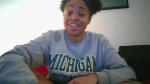 Media: A video of a smiling young African-American woman with a natural hairstyle, wearing a grey Michigan State University sweatshirt. She sits on a dark couch in a dimly lit room.