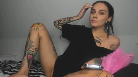 Media: Video of a tattooed woman with dark hair, wearing a black off-shoulder top and zebra-patterned shorts, sitting on a bed.