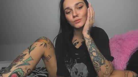 Media: Video of a tattooed woman with long black hair, sitting on a zebra-print bed, wearing a black T-shirt, with a grey wall background.