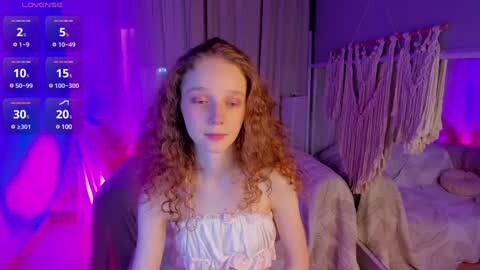 Media: A video of a young, fair-skinned girl with curly, reddish-brown hair, wearing a light-colored, lacy dress. She sits in a dimly lit bedroom with purple and pink lighting, next to a bed with a fringed blanket.