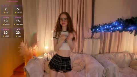 Media: Video of a slender, fair-skinned woman with long brown hair and glasses, wearing a white crop top and black plaid mini skirt, standing in a cozy living room with plush furniture, string lights, and a potted plant.