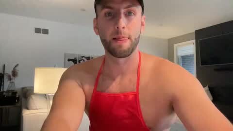 Media: Video of a muscular, bearded Caucasian man with short dark hair, wearing a red apron, standing in a modern living room with beige walls, a TV, and a lamp.