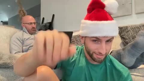 Media: Video: A man in a red Santa hat and green shirt plays a video game on a couch, while a bald man in glasses watches.
