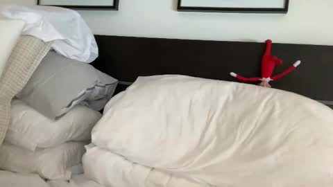 Media: Video of a messy bed with white sheets, grey pillows, and a red Elf on the Shelf toy on the headboard.