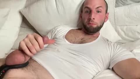 Media: Video of a muscular, light-skinned man with a shaved head and beard, wearing a tight white T-shirt, lying on a white bed, holding his erect, circumcised penis.