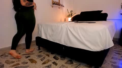 Media: A video shows a woman in black leggings and bare feet on a mosaic-tiled floor, near a bed with white sheets, black pillows, and a lit lamp.