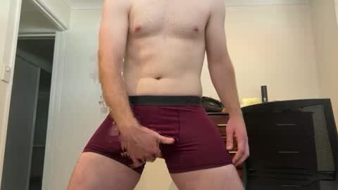 Media: Video of a light-skinned, shirtless man in maroon boxer briefs, standing indoors, holding his underwear. Background features a closed door, a dresser, and a TV.