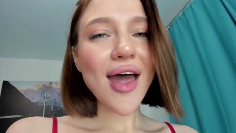 Media: Video of a young Caucasian woman with shoulder-length brown hair, fair skin, and light makeup. She has a playful expression with her tongue out, wearing a red spaghetti-strap top. Background includes a teal curtain and a blurred mountain landscape painting.