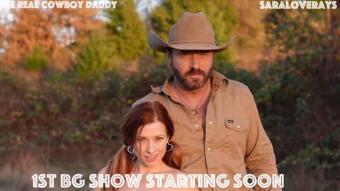 Media: Video of a rugged cowboy with a beard and a woman with long red hair, both wearing denim shirts, standing in a wooded outdoor setting. Text overlays read \"REAL COWBOY DADDY\" and \"SARAH OVEREASY.\
