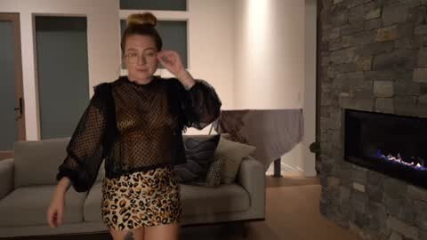 Media: A video of a blonde woman in a sheer black mesh top and leopard print skirt, adjusting her hair in a modern living room with a stone fireplace and grey couch.