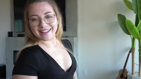 Media: Video of a smiling, fair-skinned woman with long blonde hair, wearing glasses and a black V-neck top, standing in a modern living room with a green plant and a TV in the background.