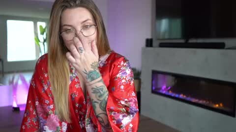 Media: Video of a fair-skinned woman with long, straight blonde hair, wearing glasses, and a floral kimono, covering her face with her hands. Background includes a modern living room with a fireplace and potted plants.