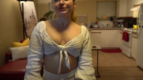 Media: Video of a fair-skinned, red-haired woman with glasses wearing a white, knotted crop top, in a modern, warmly lit kitchen with white cabinets and red accents.