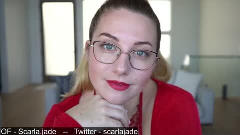 Media: Video of a fair-skinned woman with blue eyes, wearing red lipstick, glasses, and a red cardigan, smiling in a modern, brightly lit room.