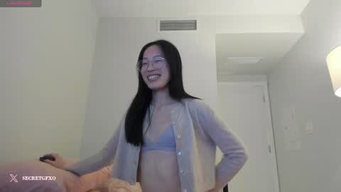 Media: A smiling Asian woman with long black hair and glasses, wearing a sheer gray cardigan over a light blue bra, is being hugged by a person in a beige shirt. The setting is a modern, white-walled room with a door in the background.