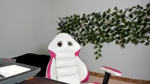 Media: A video of a modern gaming chair with white and pink accents, positioned in front of a gray wall adorned with a green, leafy, lattice-patterned wallpaper. A black laptop and a white keyboard are partially visible on the left.