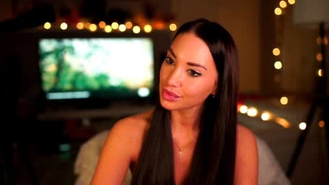 Media: Video of a smiling woman with long, straight black hair, fair skin, and light pink lipstick, sitting in a dimly lit living room with a TV screen displaying a green forest scene in the background.