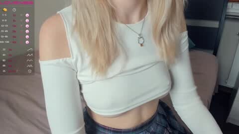 Media: Video of a young woman with long blonde hair, wearing a white off-shoulder crop top and high-waisted jeans, sitting on a bed. Background shows a smartphone screen with app icons.