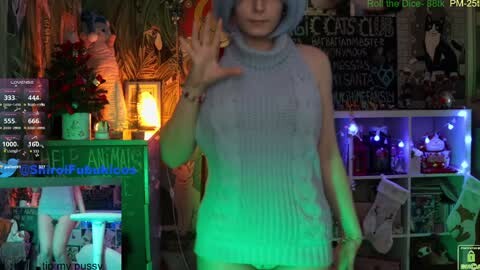 Media: Video of a woman with short blue hair in a grey knitted sweater, taking a selfie in a dimly lit, cluttered room with various decorations, including a lit candle and a green screen.