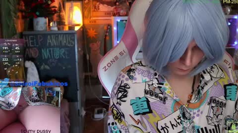 Media: Video of a pale-skinned woman with blue hair, wearing a multicolored, cartoon-stickered hoodie, standing indoors. Background features a TV, \"Zoome\" sign, and a chalkboard with \"LEARN ANIMALS AND NATURE\" in chalk.