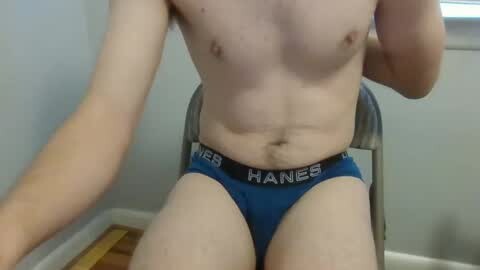 Media: Video of a fair-skinned, shirtless man with a lean physique, wearing blue Hanes briefs, sitting on a wooden chair against a plain gray wall.