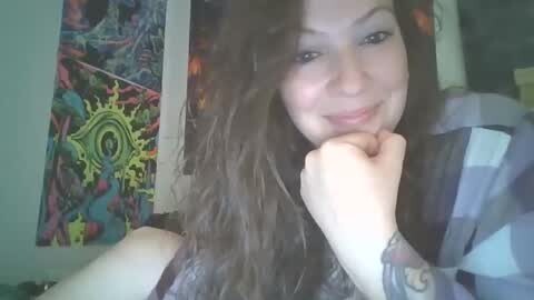 Media: Video of a smiling, curly-haired woman with fair skin, wearing a plaid shirt, sitting on a bed with a colorful, psychedelic tapestry in the background.