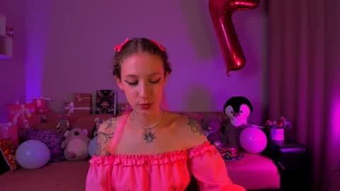 Media: A video of a young woman with light skin, dark hair in pigtails, and tattoos, wearing a pink off-shoulder top, standing in a dimly lit room with a red balloon, stuffed toys, and a \"7\" sign.
