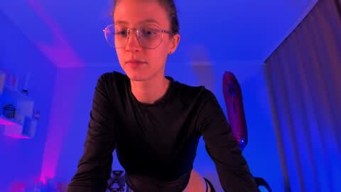 Media: Video of a young woman with fair skin and glasses, wearing a black long-sleeve top, against a purple-lit background.