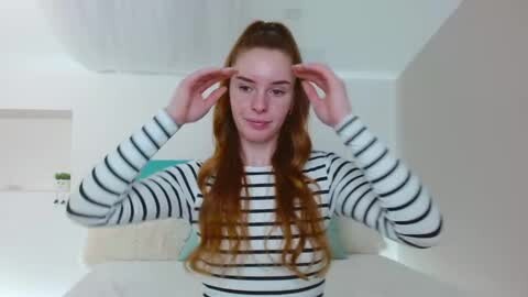 Media: Video of a young woman with long, wavy red hair, fair skin, and blue eyes, wearing a black and white striped long-sleeve shirt, adjusting her hair in a modern, minimalist bathroom with white walls and a ceiling light.