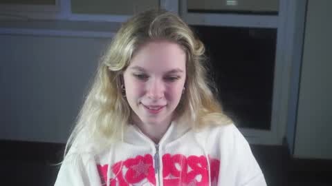 Media: Video of a young blonde woman with fair skin, wearing a white hoodie with \"KILLER\" in red letters, smiling, in a dimly lit room with white window frames.