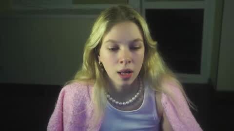 Media: Video of a blonde girl with fair skin, wearing a light pink, glittery cardigan over a white tank top and a pearl necklace, indoors with a dark background.
